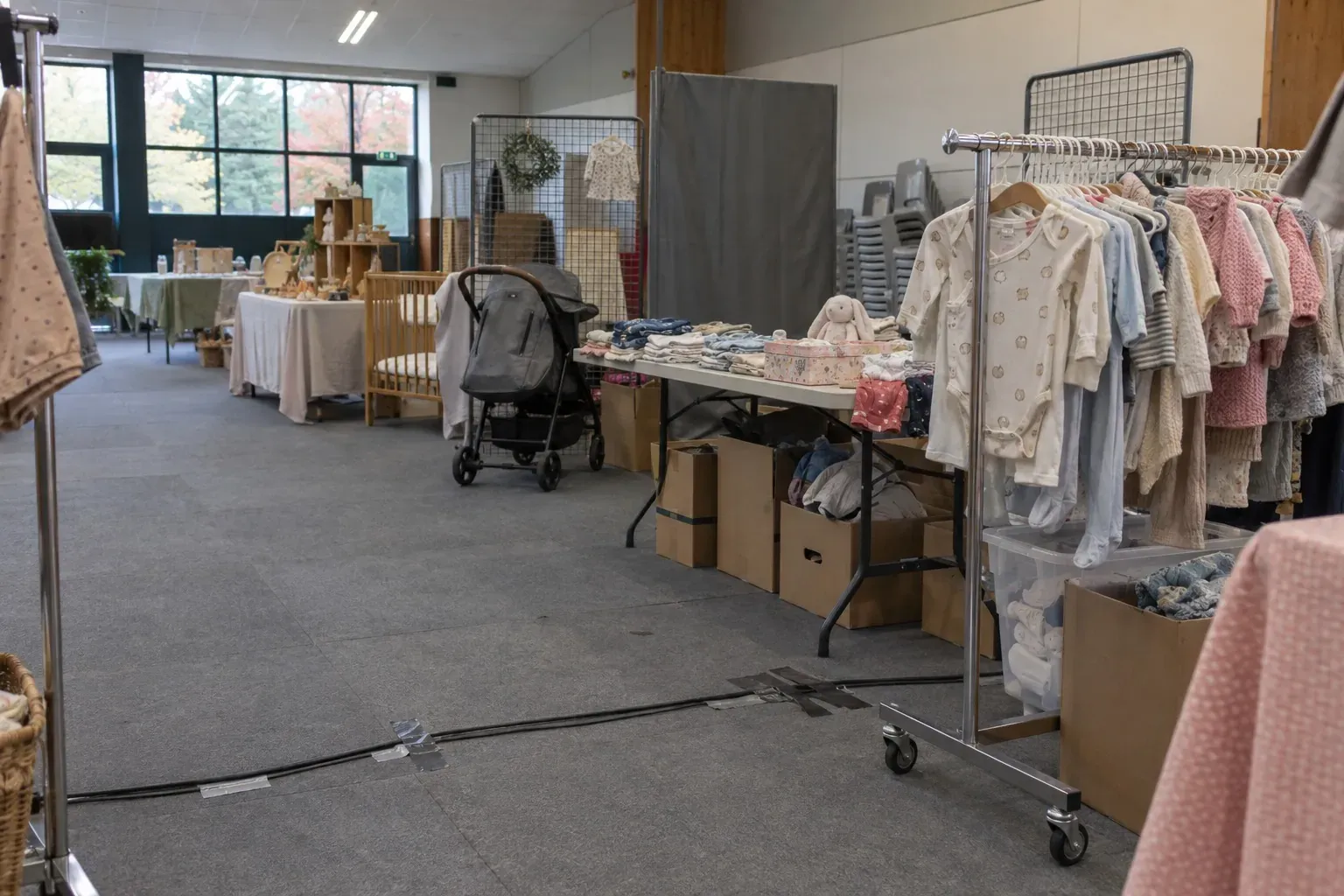 Portants de vêtements de bébé pliés sur un stand associatif, salle vide, lumière naturelle
