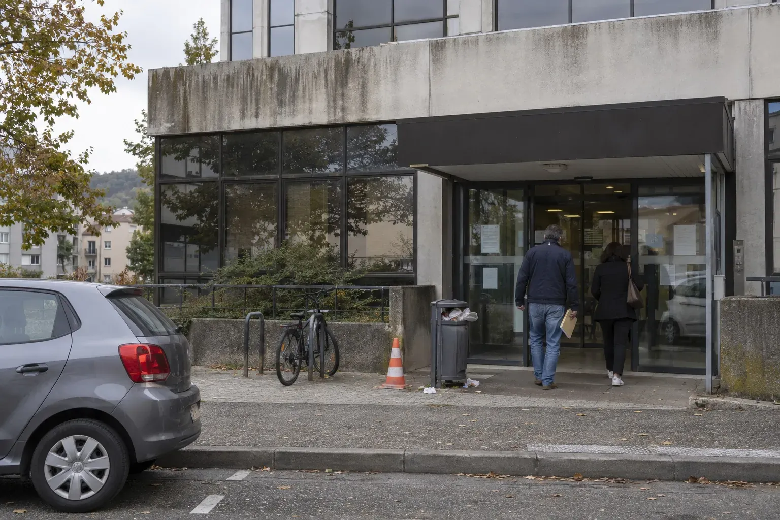Façade d'une agence d'allocations familiales en béton et verre, avec quelques adultes de dos devant l'entrée par ciel voilé