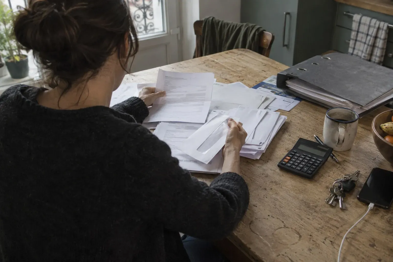 Mains d'adulte triant des courriers et formulaires administratifs sur une table de cuisine, avec calculatrice et tasse de café
